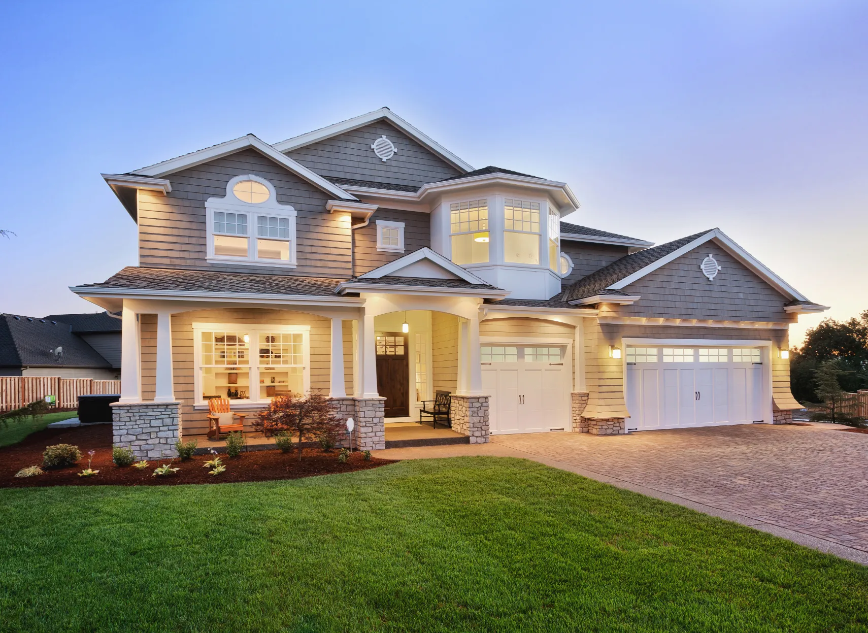 A stately two-story suburban house at dusk, its exterior lights aglow and perfectly manicured lawn framing the inviting front entrance.